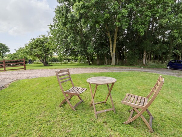 An outdoor seating area with a round table and two wooden chairs, surrounded by lush green grass and trees.