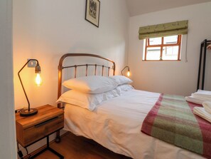 A single bed with a metal headboard, a bedside table with a lamp, a window with a green valance, and a framed picture on the wall. - The Stables (Preston)