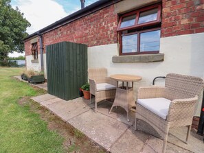 A patio with wicker furniture and a small table, adjacent to a brick building with a window. - The Stables (Preston)