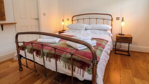 Una cama bien hecha con sábanas blancas y una manta escocesa, dos lámparas de noche, una mesita de noche de madera y una puerta a la izquierda.