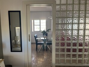 Un comedor con mesa de vidrio, sillas y una planta en maceta. Un acceso da a una habitación con suelo de baldosas y ventana.