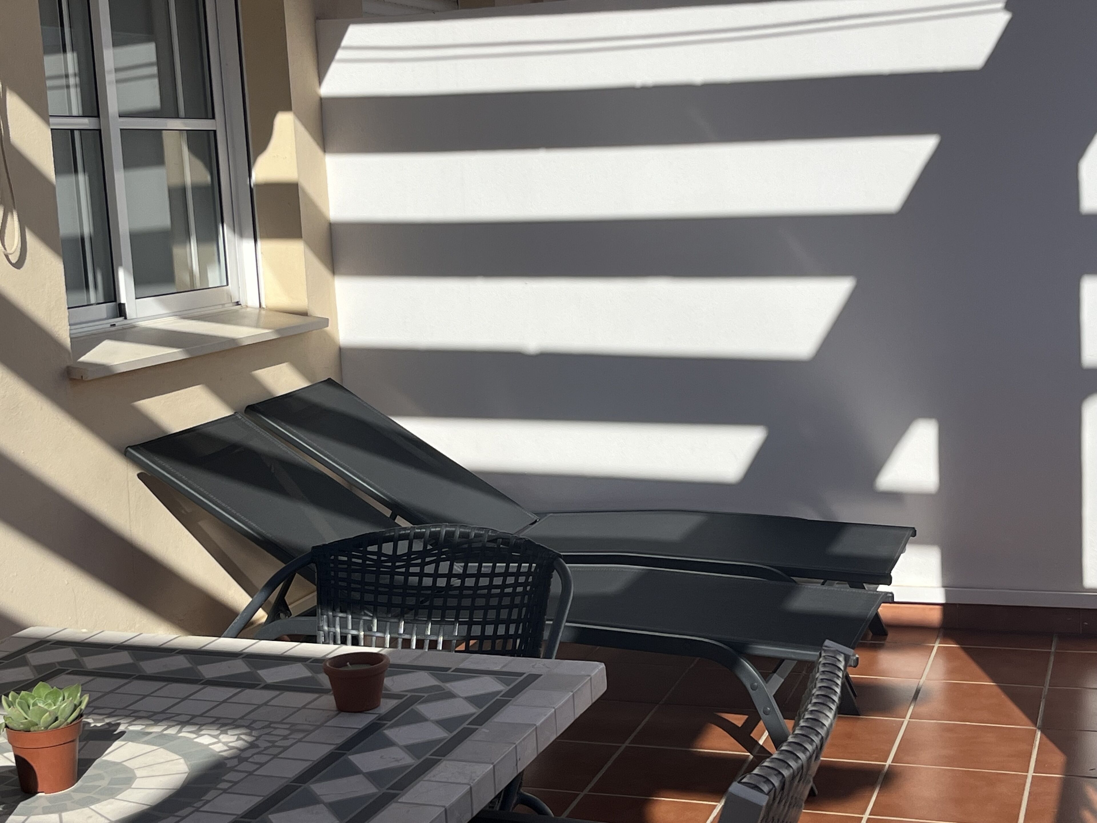 Une terrasse ensoleillée avec une table, deux fauteuils, une plante en pot et une fenêtre.