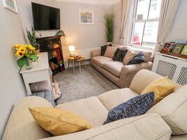 Una sala de estar con un sofá color beige, una mesita con una lámpara, una chimenea, un televisor montado en la pared y una ventana con cortinas.