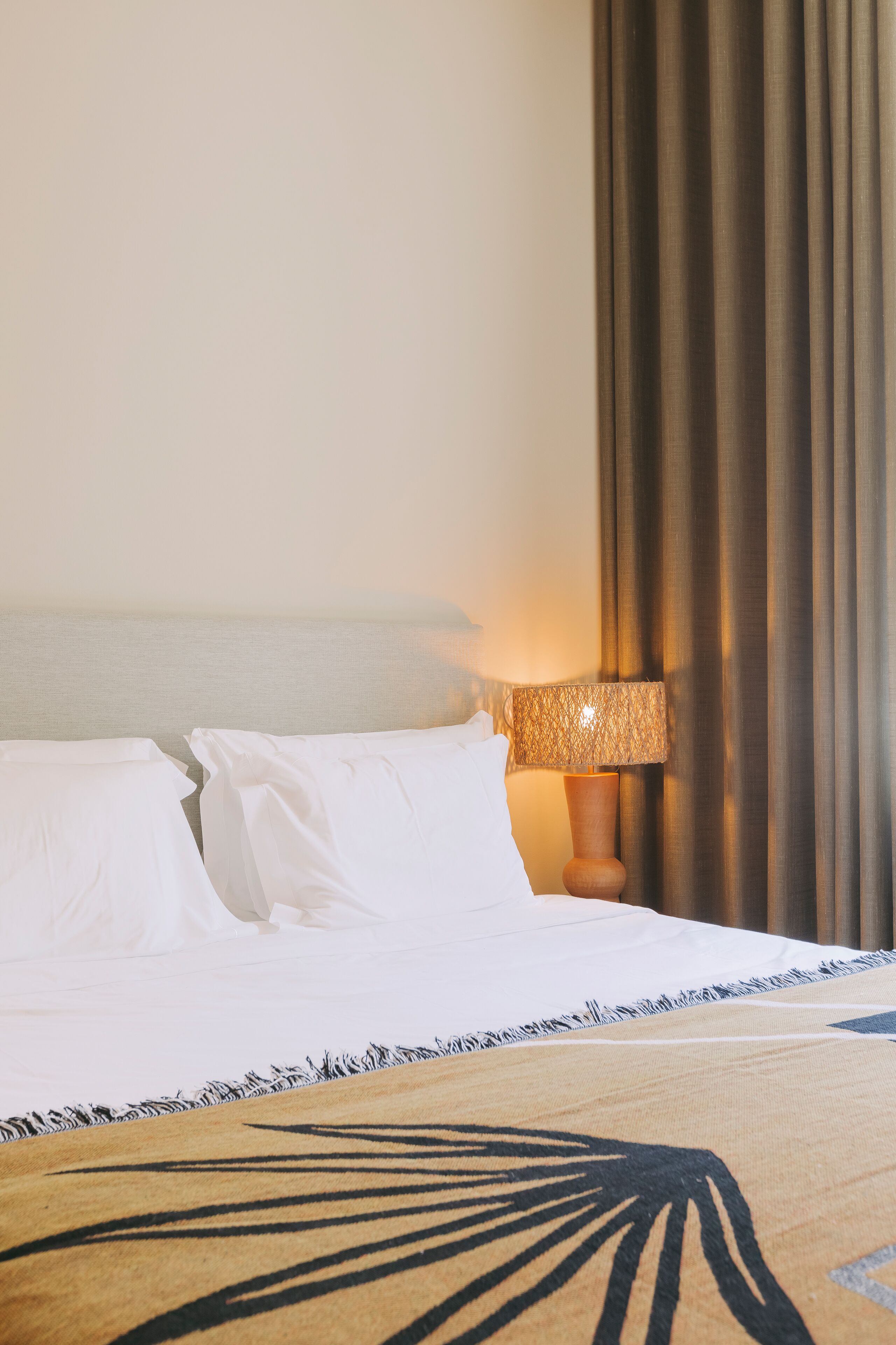 A neatly made bed with white linens and a patterned throw, a wooden bedside table with a lamp, and a view of a curtain.