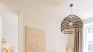 A modern hotel room with a wooden floor, a large bed, a television, a wall-mounted artwork, a hanging light fixture, and a view of the outside through a window. - Village Aparthotel by BOA (Porto)