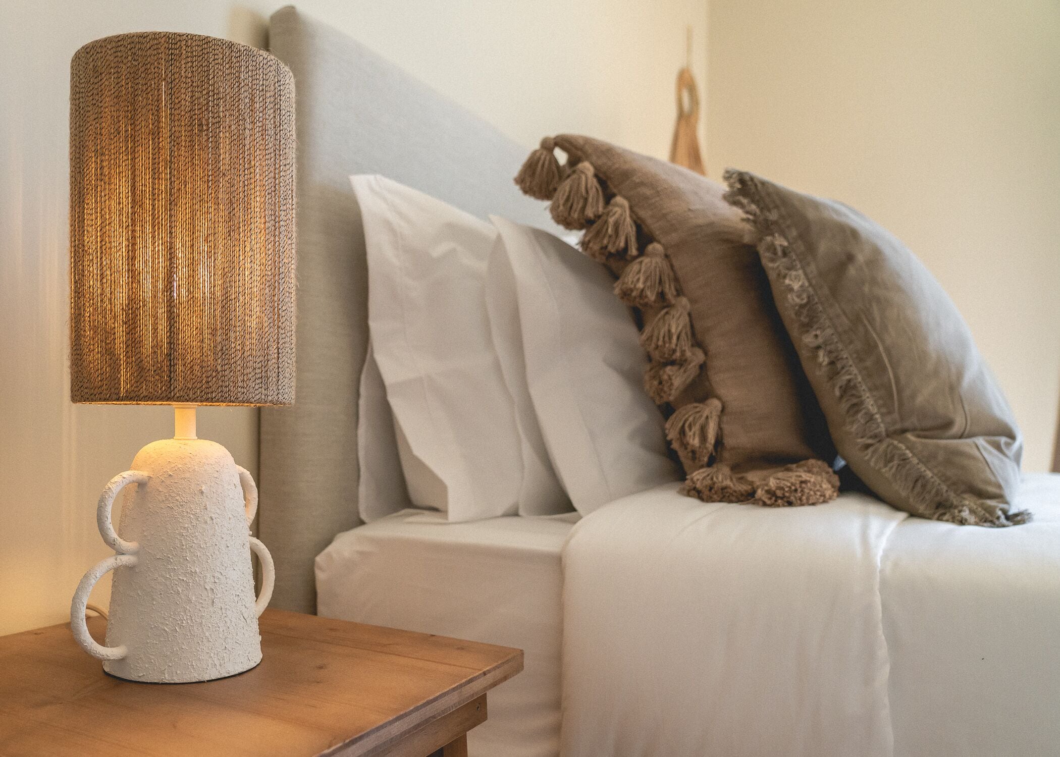 Un dormitori acollidor amb un llit blanc, una tauleta de nit de fusta, una làmpada amb textura i una manta decorativa.