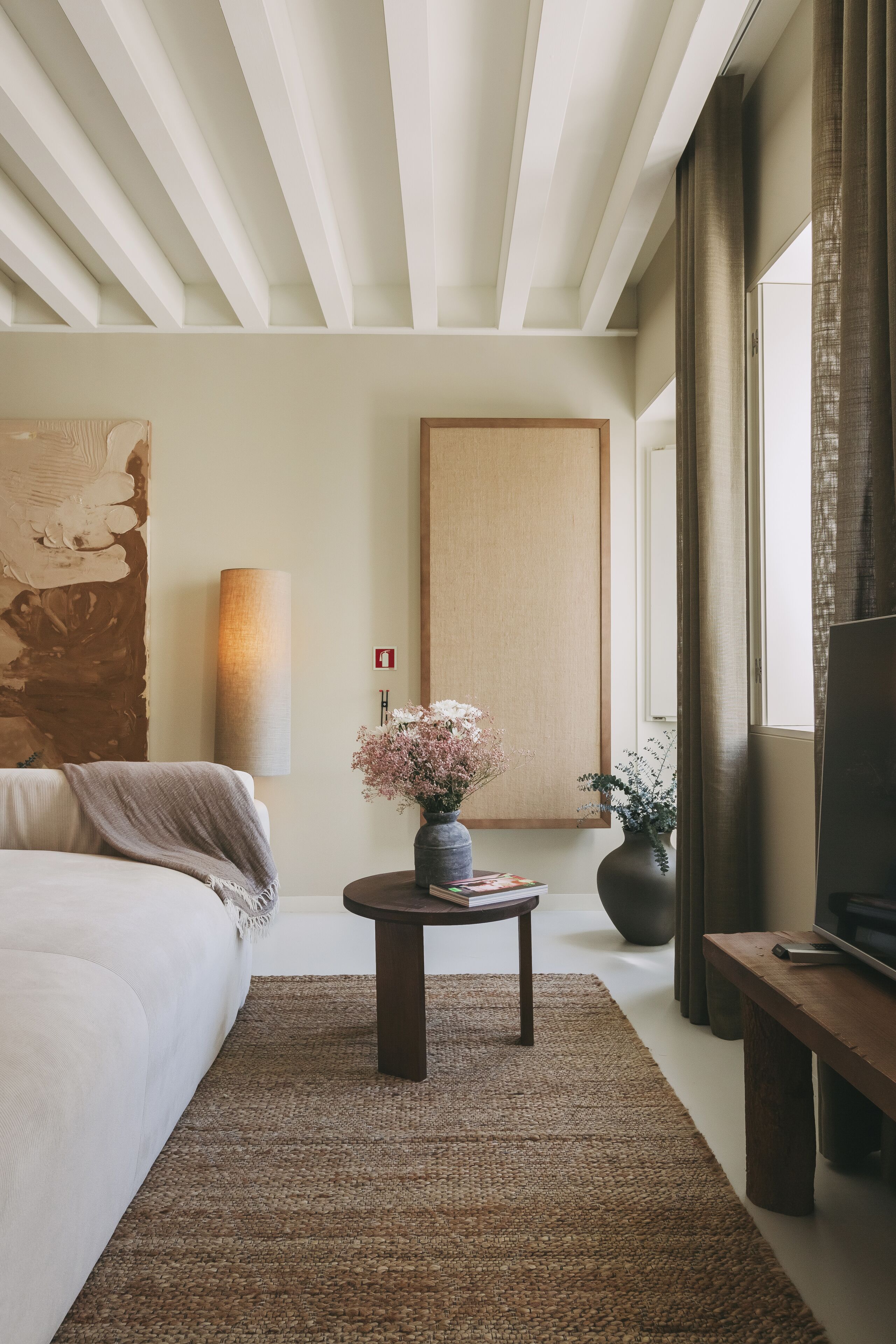 Une chambre d’hôtel moderne avec un grand lit, une table basse ronde ornée de fleurs, un grand tableau au mur et un téléviseur posé sur un meuble en bois.