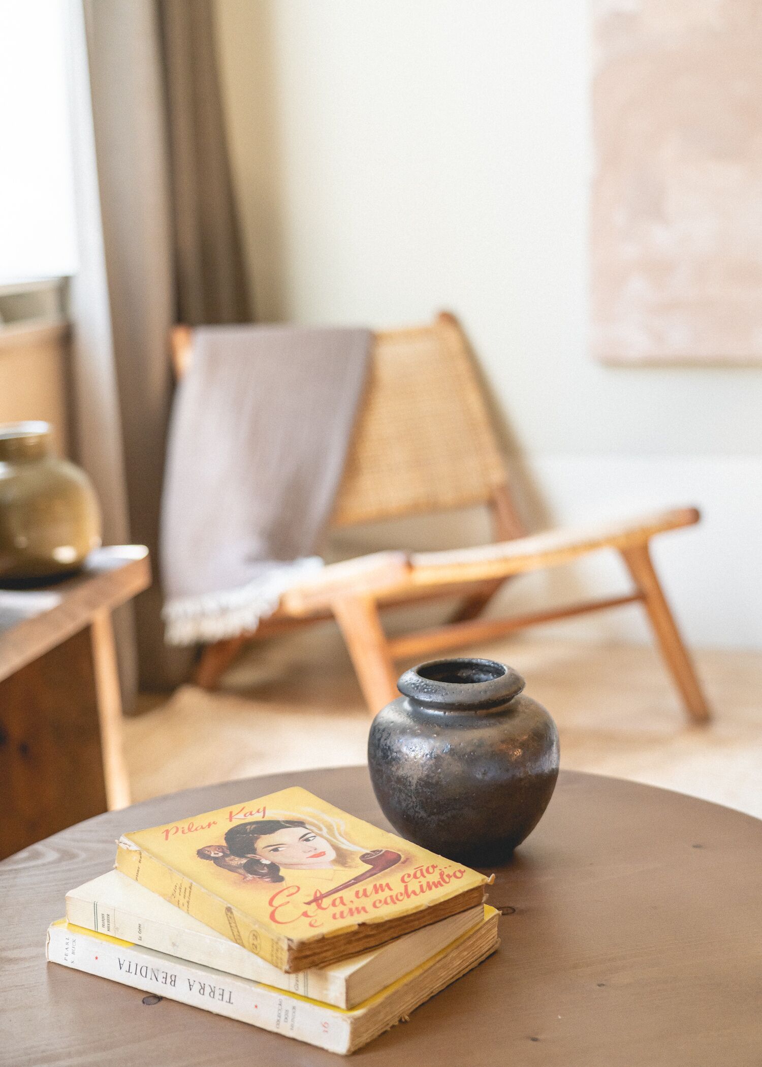 A wooden chair with a cushion, a small table with books and a ceramic vase, and a window with curtains.