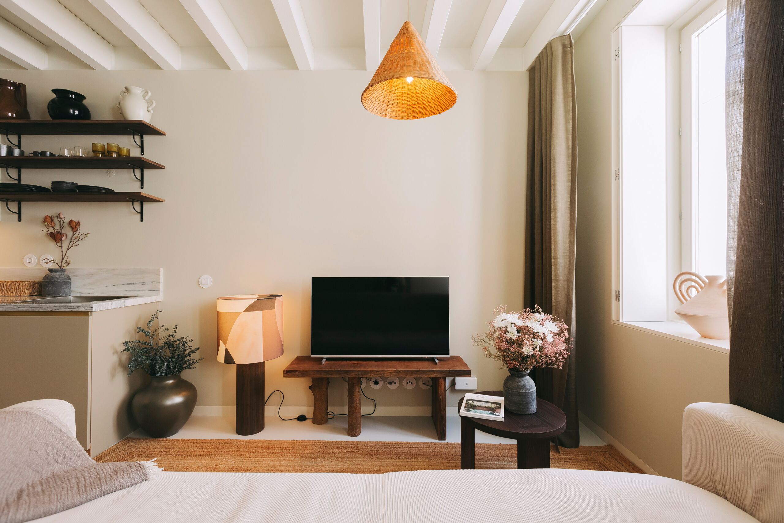 A modern hotel room with a flat-screen TV on a wooden stand, a large window with curtains, a bedside table with a lamp, and a bed with a white comforter.