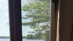 Vista a través de una ventana con marco de madera y mosquitera, sobre un lago tranquilo con un muelle y una zona de césped.