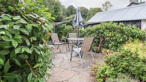 Un patio de piedra con mobiliario exterior, rodeado de vegetación exuberante y una estructura de techo metálico.