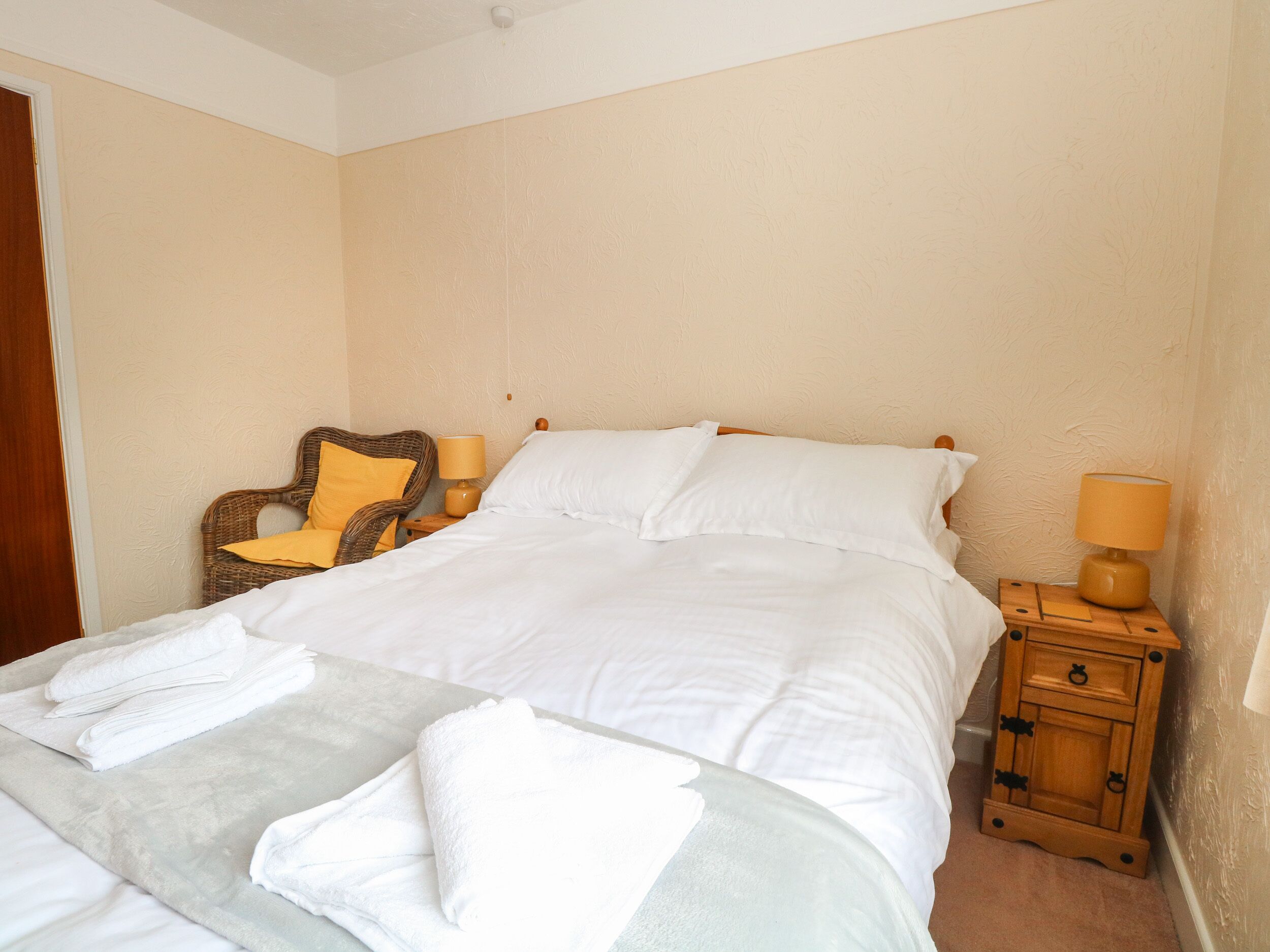 Een net opgemaakt bed met witte lakens, een houten nachtkastje met een lamp, en een rieten stoel met een geel kussen.