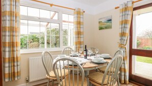 A dining room with a wooden table set for four, white chairs, and a view of the garden through large windows. - Dove's Nest (Great Yarmouth)