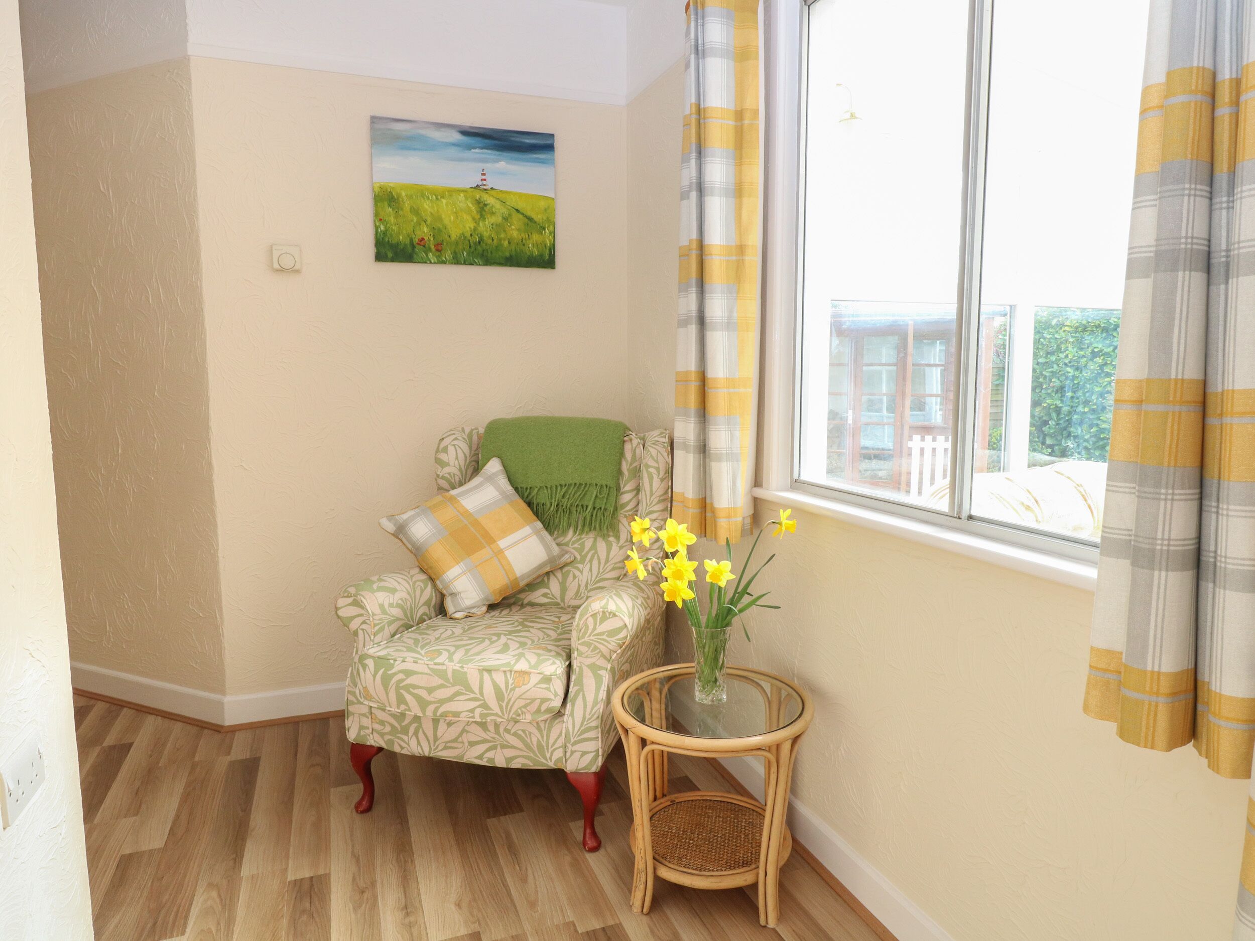 A room with a patterned armchair, a small table with a vase of flowers, a painting on the wall, and a window with curtains.