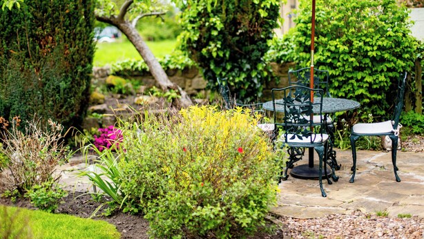 Un patio con una mesa y sillas bajo un paraguas blanco, rodeado de abundante vegetación y un camino de piedra.
