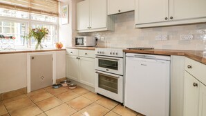 A kitchen with white cabinets, a microwave, a stove, and a dishwasher. There are pet bowls on the floor and a window with blinds. - Blue Crab Cottage (St Austell)