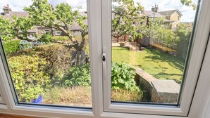 Uitzicht door een raam op een tuin met een houten terras, een stenen muur en verschillende planten en bomen.