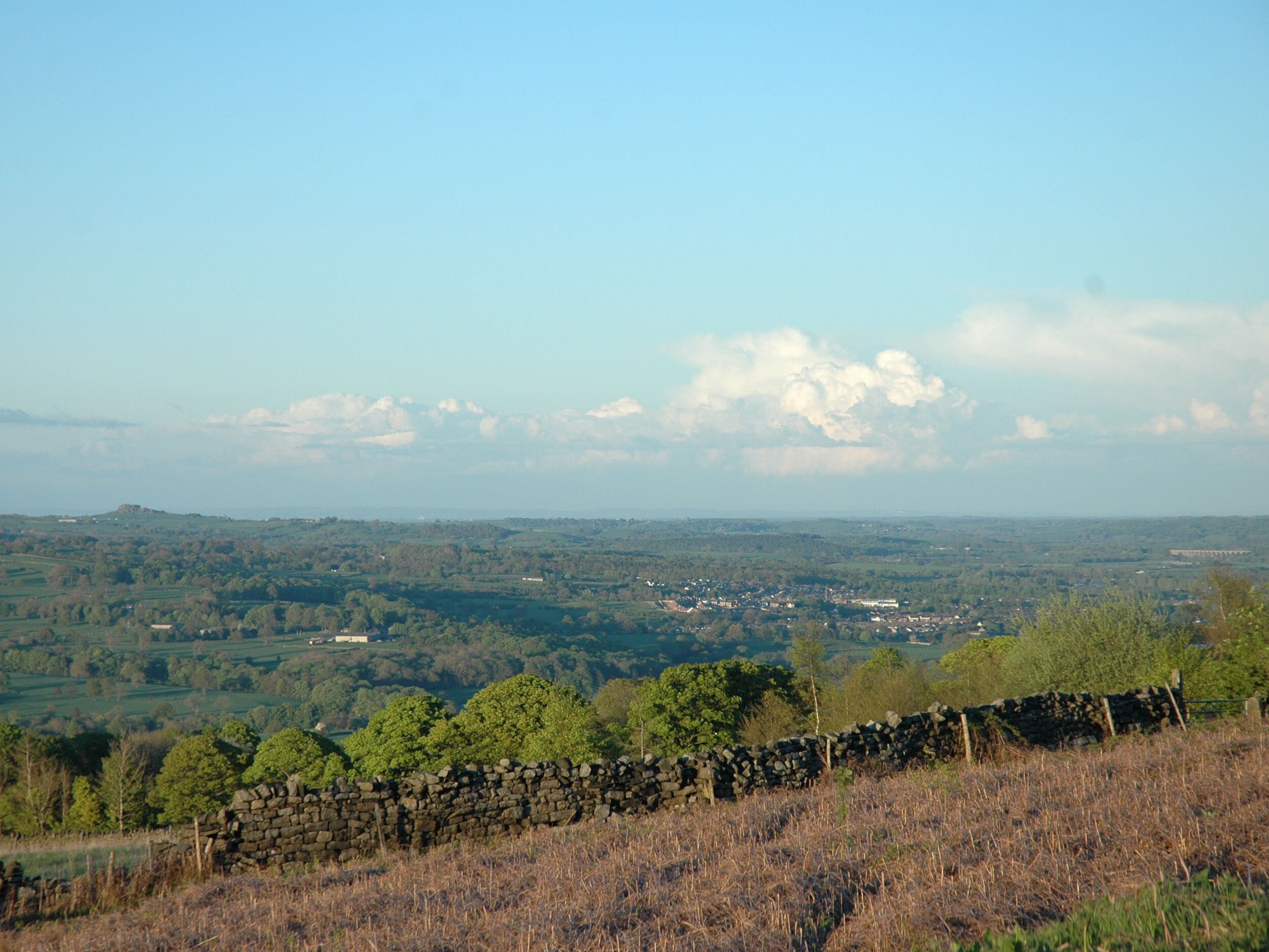 A rural landscape with a stone wall in the foreground, a small town in the midground, and a vast expanse of greenery and hills in the background.