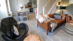 Een moderne woonkamer met een bruine leren bank, een zwarte fauteuil en een eethoek met een witte tafel en stoelen. Een trap leidt naar een hoger gelegen verdieping.