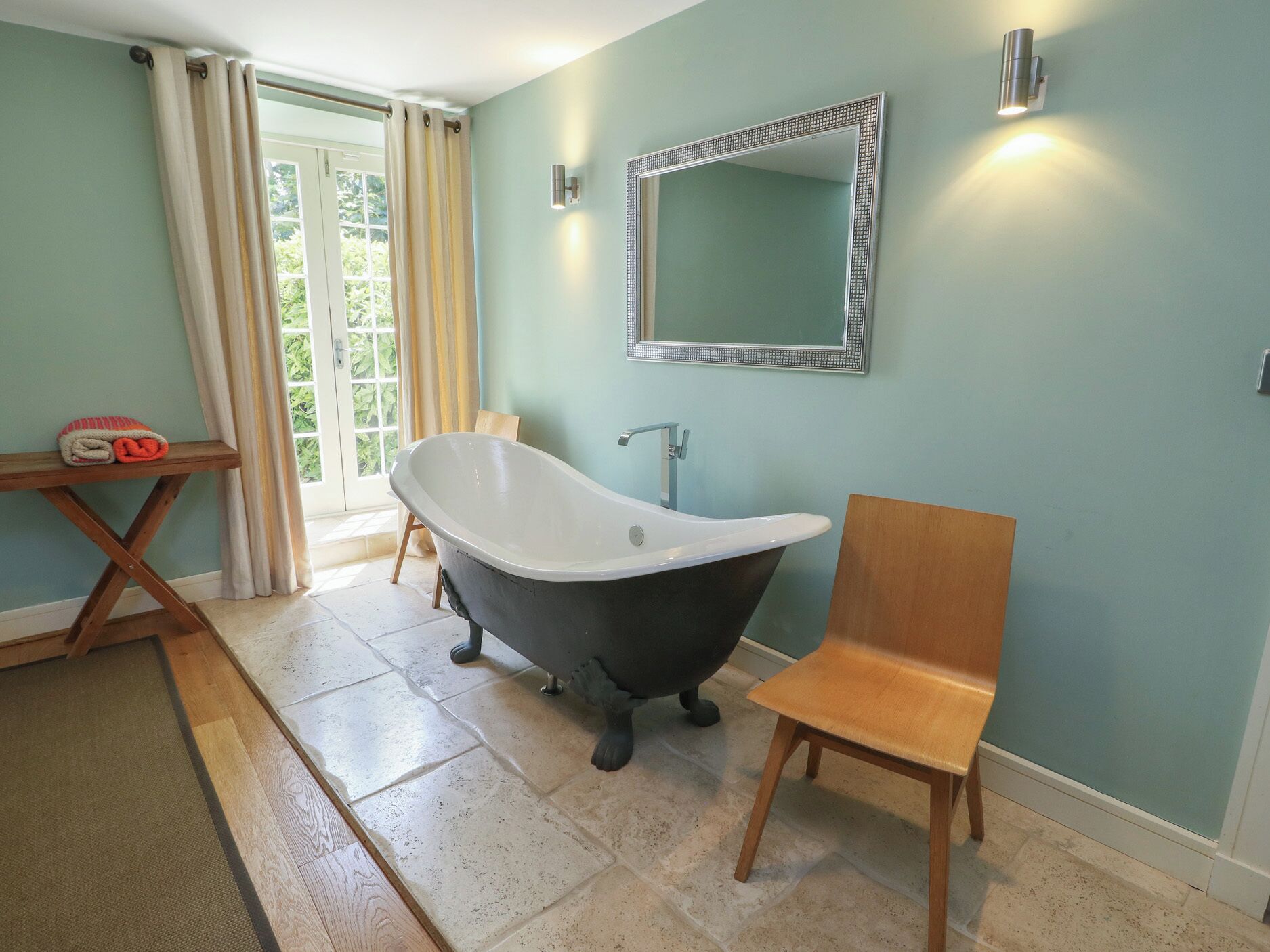 Une salle de bain avec une baignoire sur pieds, une chaise en bois, un miroir et une fenêtre avec des rideaux.