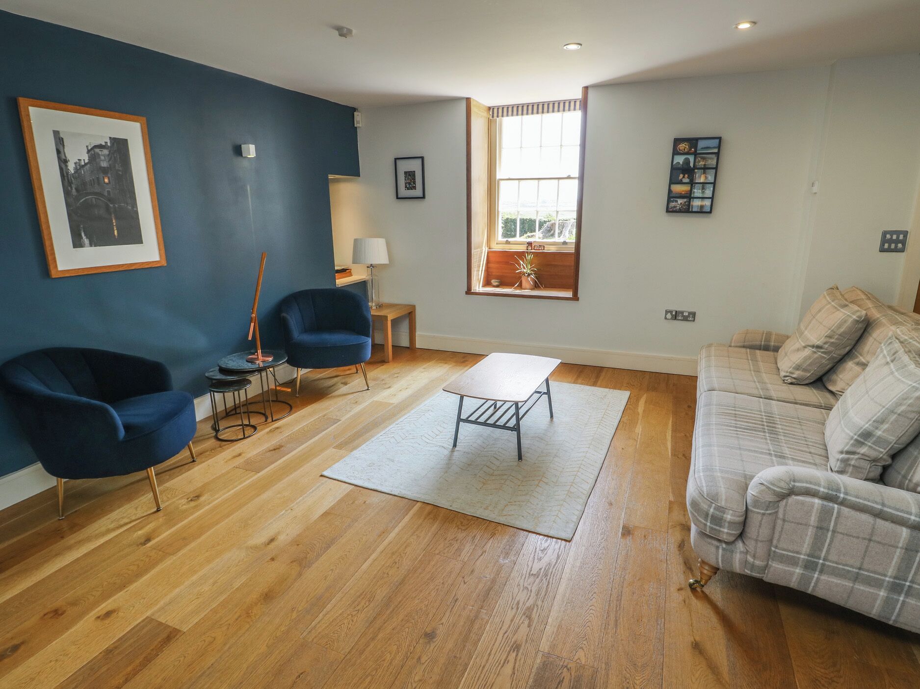 A living room with a blue wall, a wooden floor, a blue armchair, a grey sofa, a small table, and a window with a view.