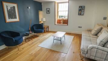 A living room with a blue wall, a wooden floor, a blue armchair, a grey sofa, a small table, and a window with a view.
