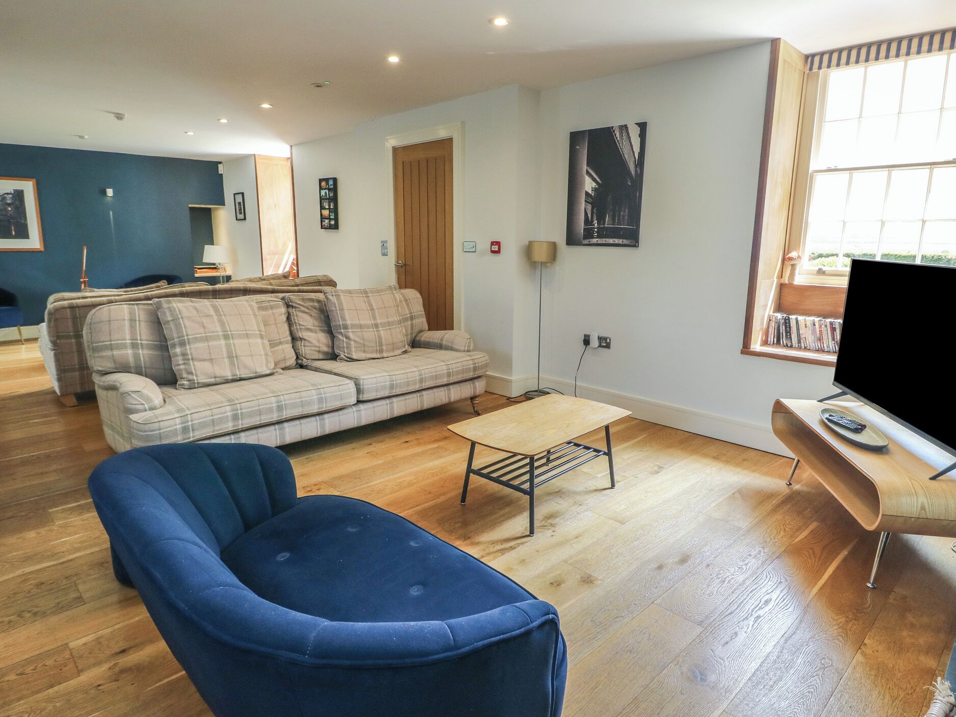 Un salon avec un fauteuil bleu, un canapé à carreaux, une table basse en bois et un téléviseur posé sur un meuble.