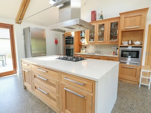 A modern kitchen with a large central island, stainless steel appliances, wooden cabinets, and a glass door pantry.