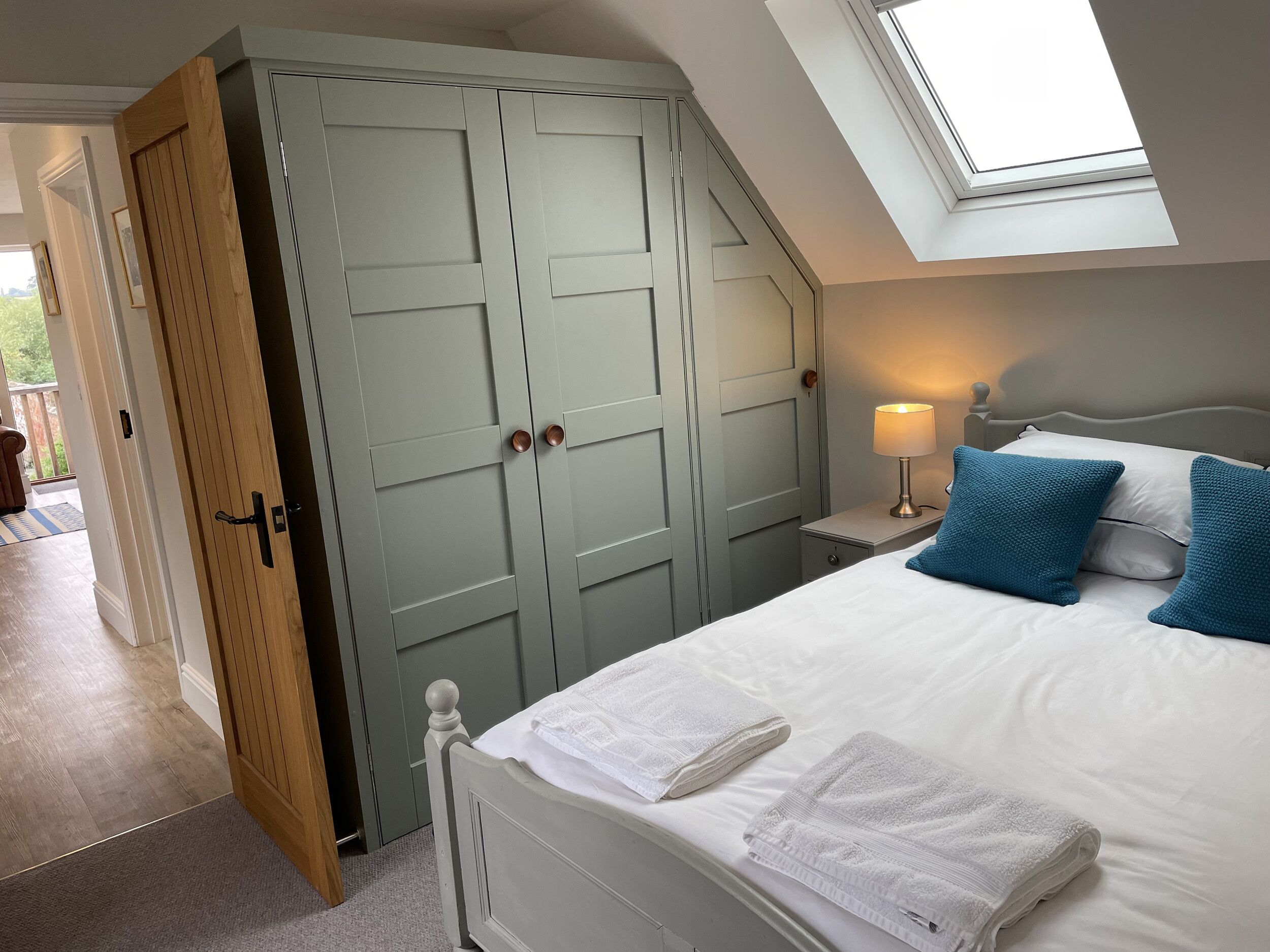 A bedroom with a skylight, a bed with white linens and blue pillows, a nightstand with a lamp, and a wardrobe with a wooden door.