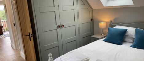 Una camera da letto con un lucernario, un letto con lenzuola bianche e cuscini blu, un comodino con una lampada e un armadio con porta in legno.