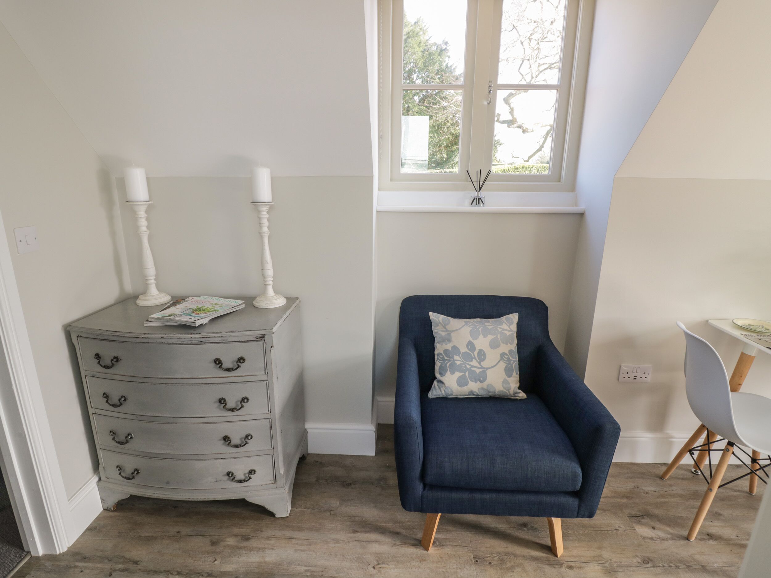 A room with a white dresser with drawers, a blue armchair with a floral pillow, and a window with a view of trees.
