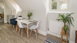 A dining area with a white table set for four, white chairs, a potted plant, and a view of the outdoors through a window. - Coach House Mews (Stratford-upon-Avon)