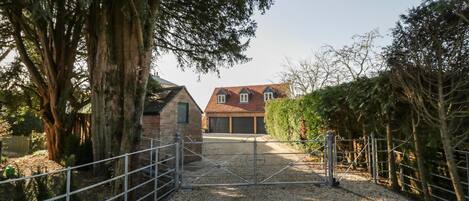 Ein Schotterweg führt zu einem Haus mit einem Metalltor, umgeben von Bäumen und einem Hecken.