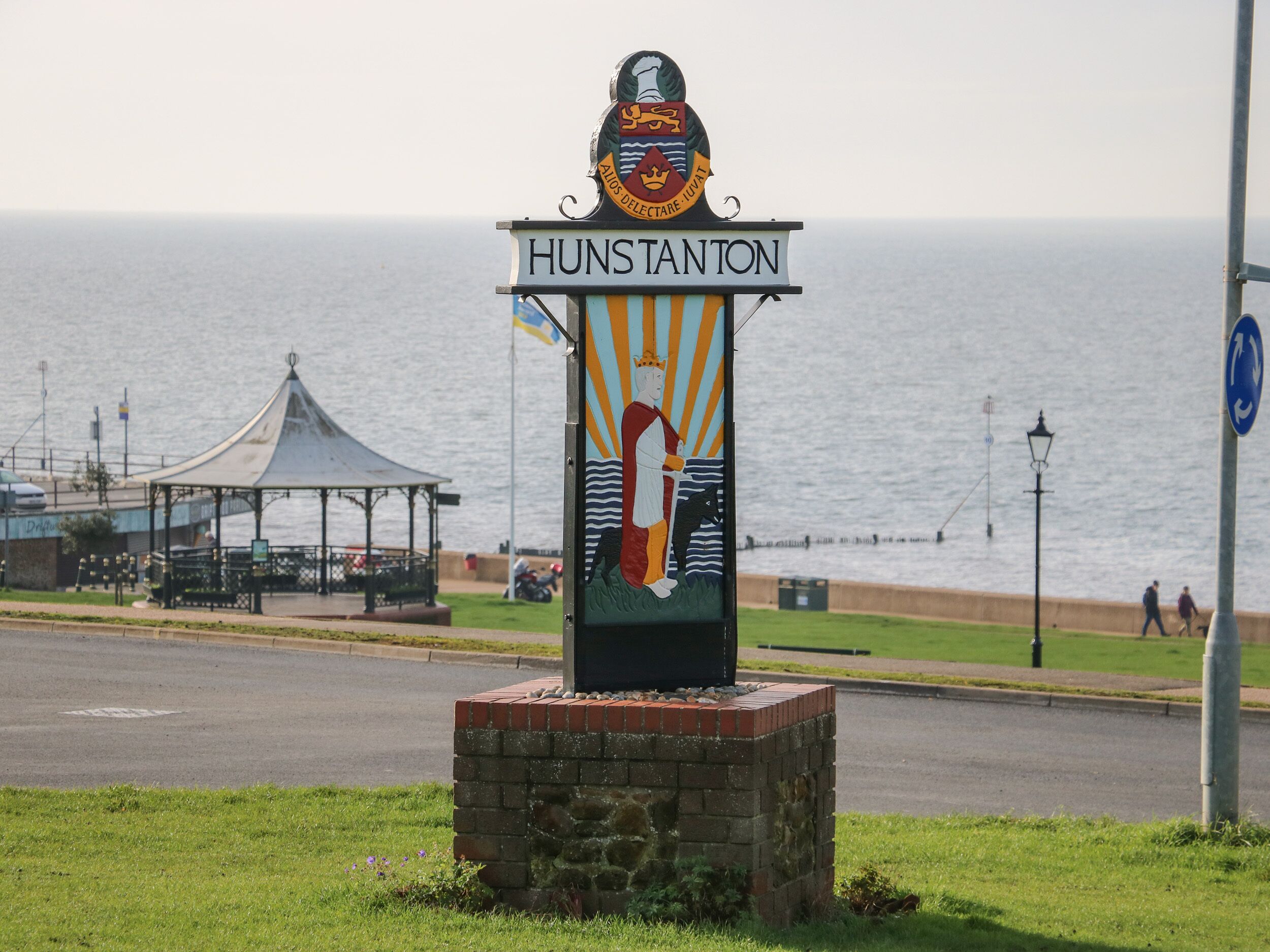 Un panneau portant l’inscription « HUNSTANTON » et affichant des armoiries, situé près d’une promenade en bord de mer, à proximité d’un kiosque et d’une route.