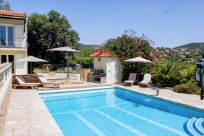 A swimming pool with lounge chairs and umbrellas, a barbecue area, and a view of a hillside.