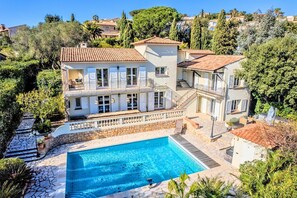 A large two-story house with a swimming pool, surrounded by trees and a hilly landscape.