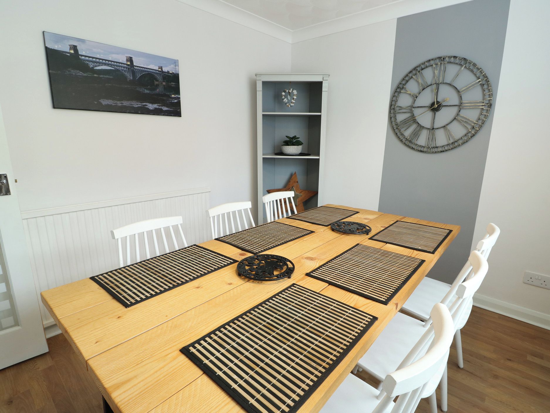 A dining area with a long wooden table, black placemats, white chairs, a decorative clock, and a framed picture on the wall.