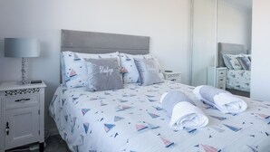 A neatly made bed with a sailboat pattern, a bedside table with a lamp, and a mirror on the wall. - Glasfryn (Holyhead)