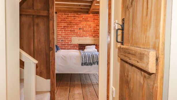 A narrow hallway leading to a bedroom with a brick wall, wooden ceiling beams, and a bed with a striped blanket. - Chevin View (Belper)