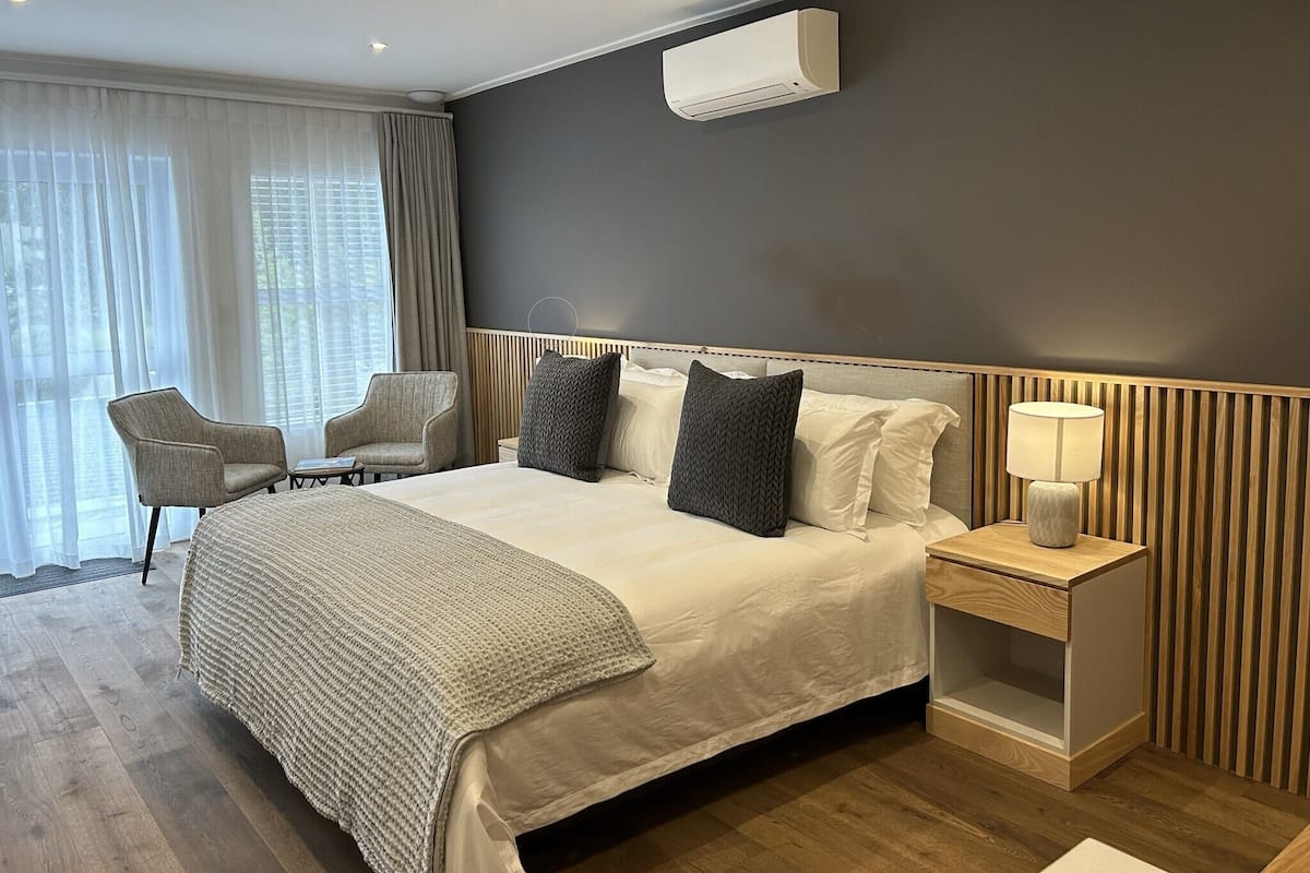 A modern hotel room with a large bed, a bedside table with a lamp, a chair, and a window with curtains.