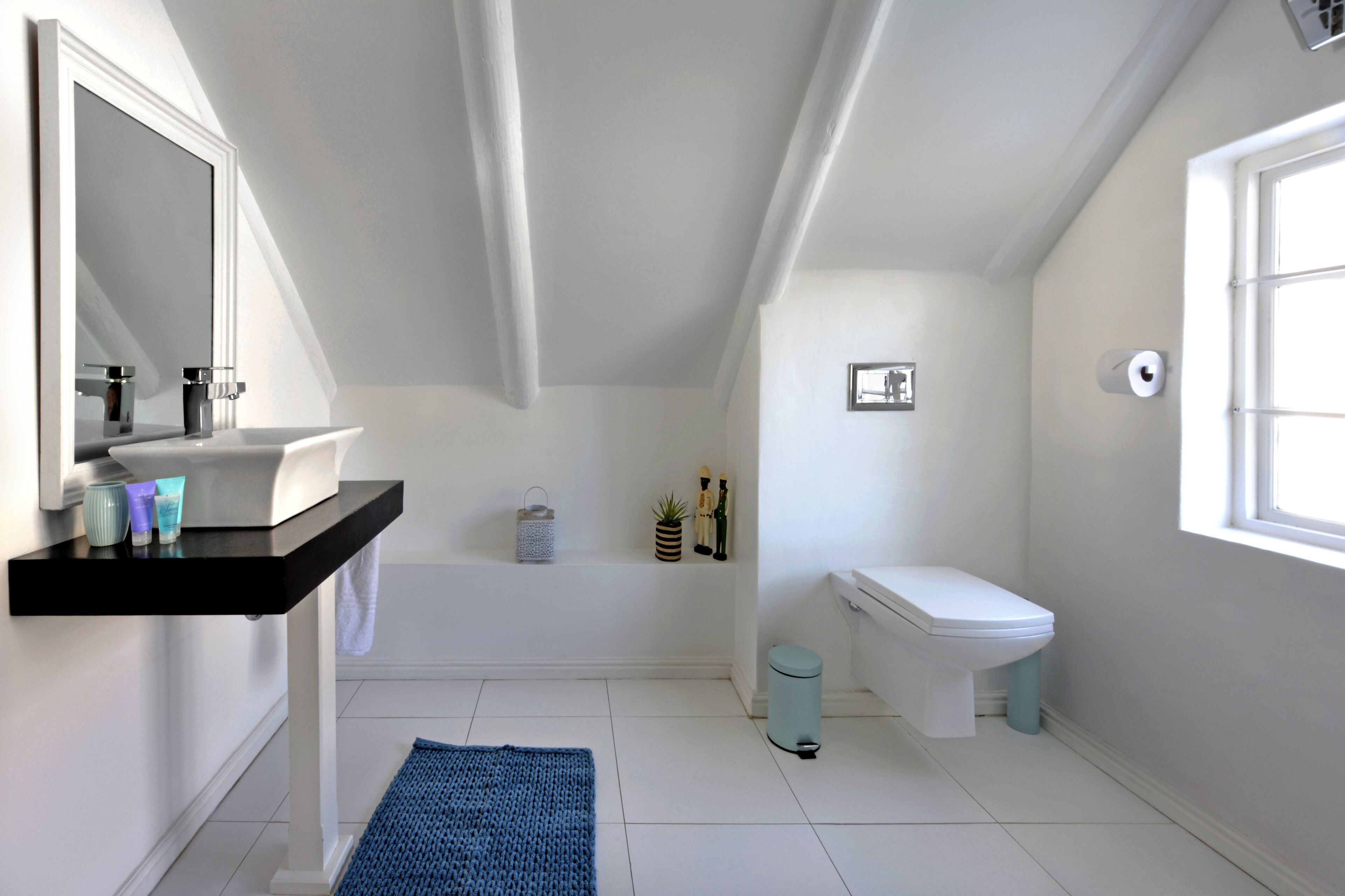 A modern bathroom with a white sink, mirror, and toilet. There is a small window providing natural light and a blue rug on the floor.