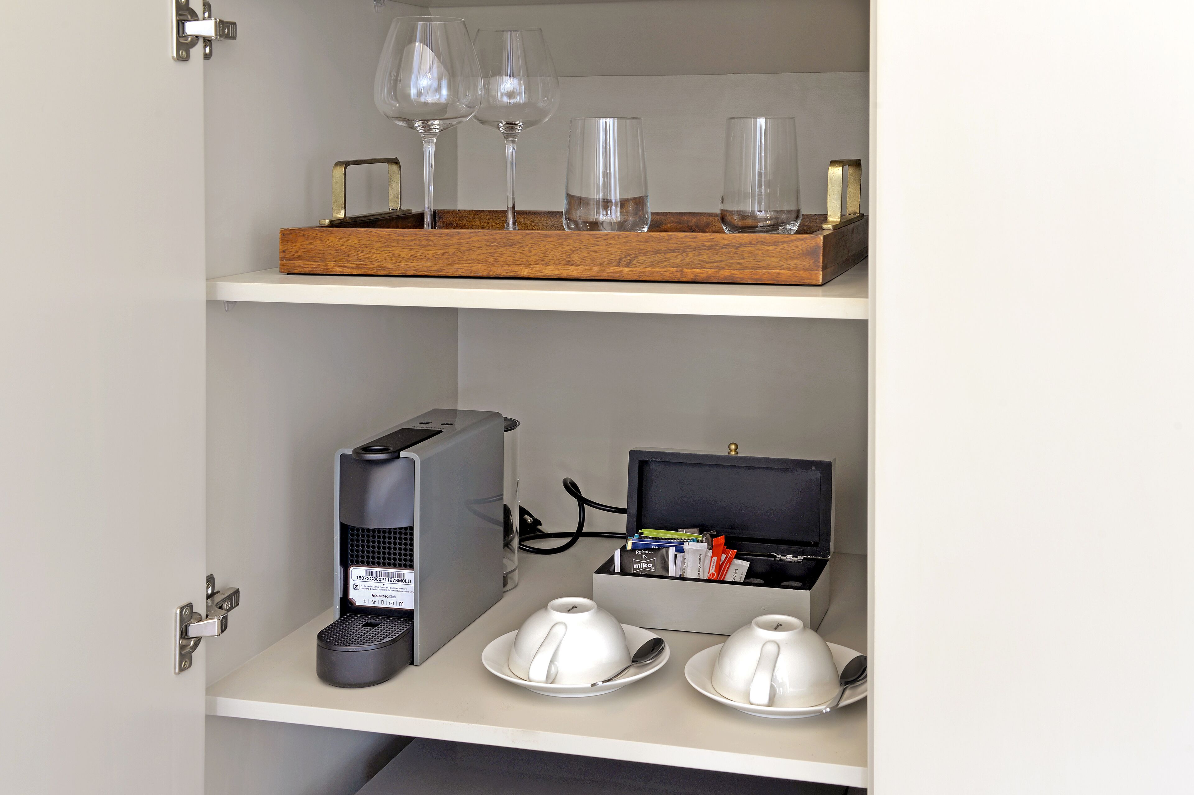 Un minibar con una cafetera, dos tazas blancas, una bandeja con copas de vino y una pequeña caja negra con objetos.