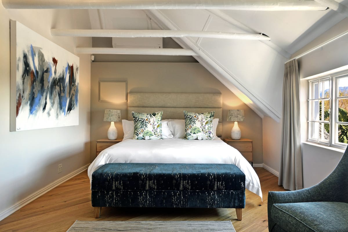 A bedroom with a large bed, two bedside tables with lamps, a dark blue bench, a large abstract painting, and a window with curtains.