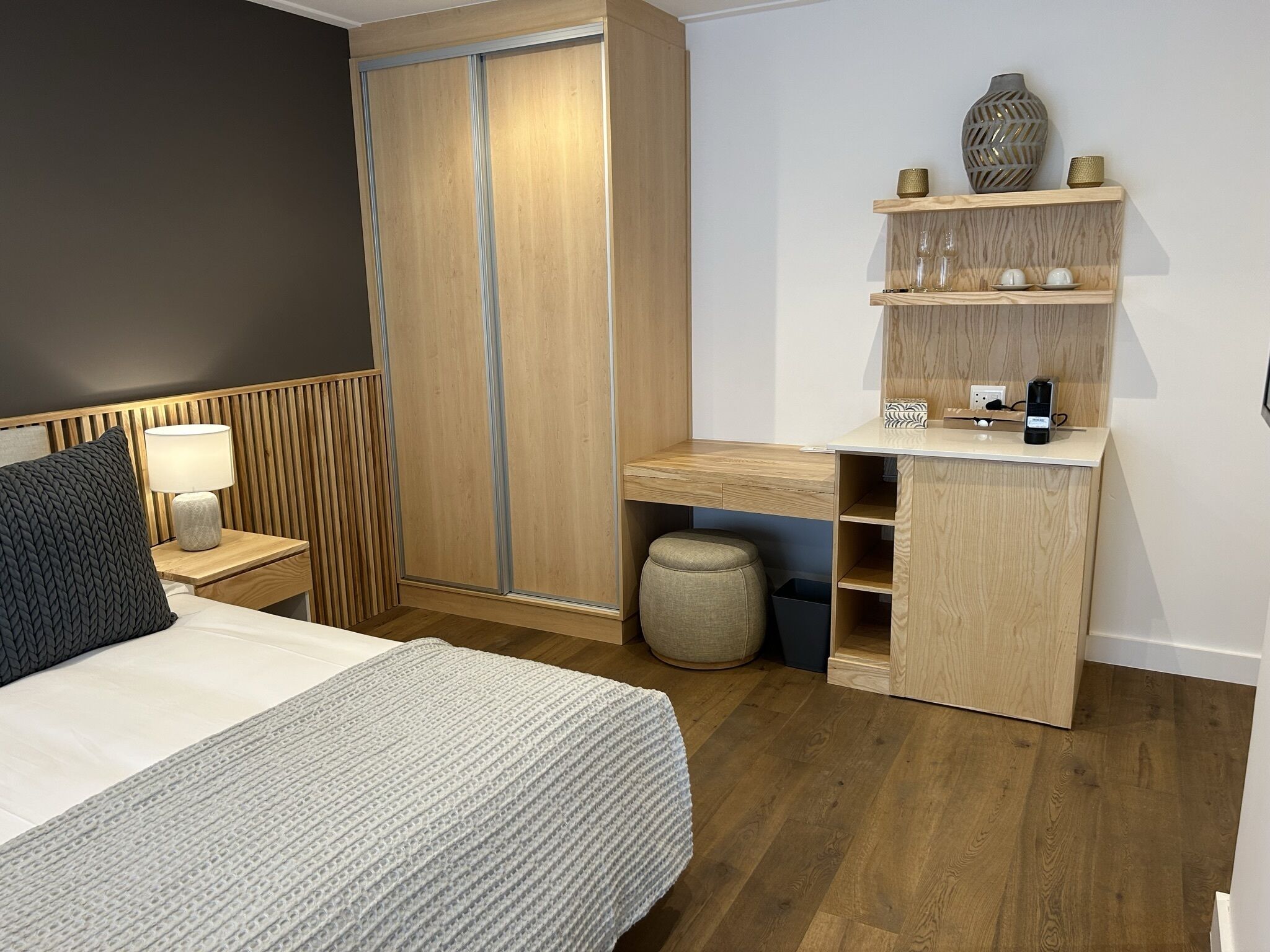 A modern bedroom with a bed, bedside table, lamp, wardrobe, and built-in desk with shelves.