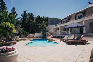 A pool area with lounge chairs and umbrellas, surrounded by potted plants and a building with a white facade and a thatched roof. - Mirabelle Guesthouse (Franschhoek)