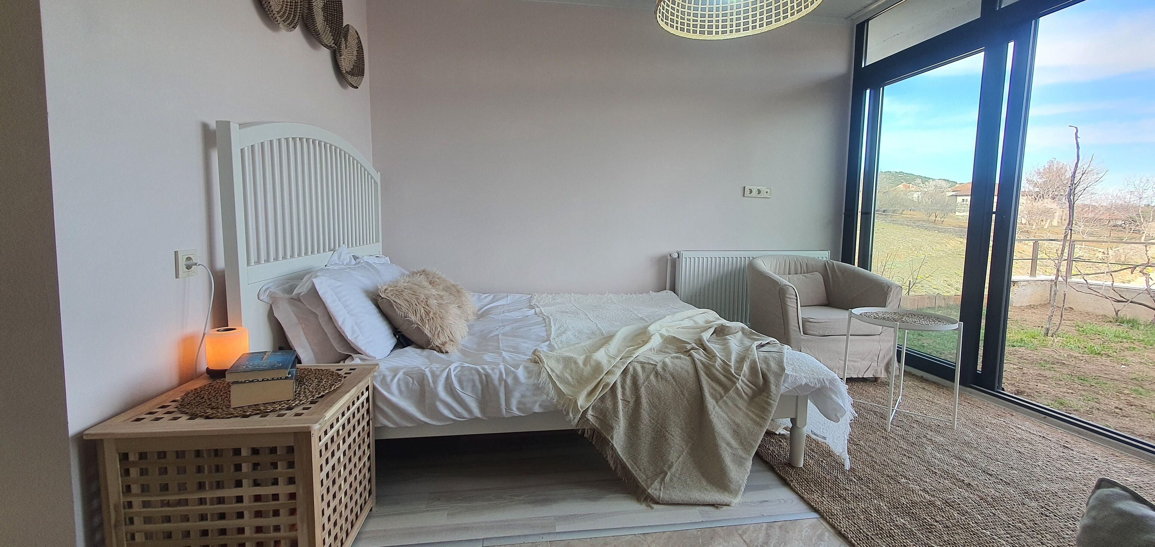 A bedroom with a bed, bedside table, lamp, and a view of the outdoors.