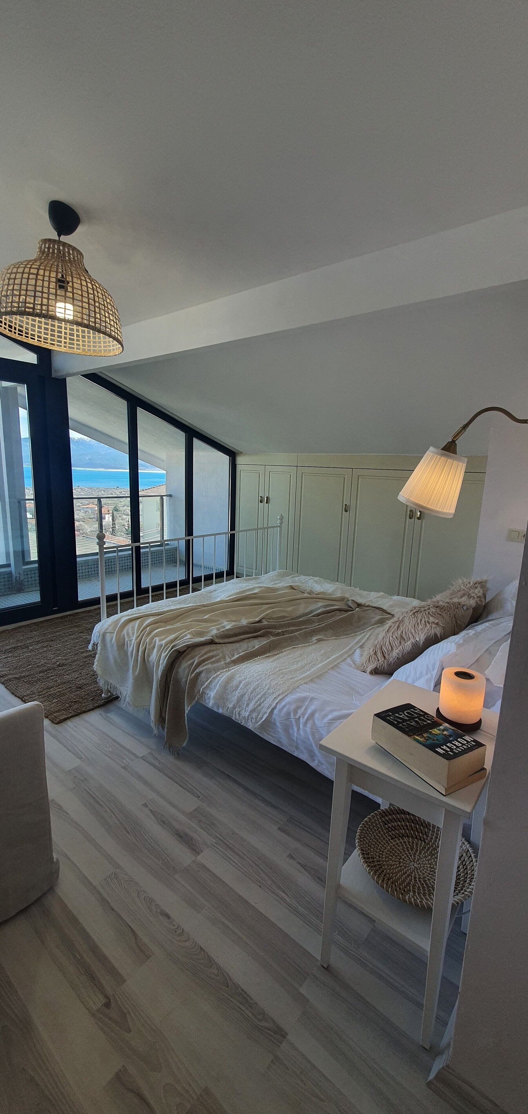 Une chambre avec un grand lit, une table de chevet avec une lampe, une vue sur la mer par de grandes fenêtres et un balcon.