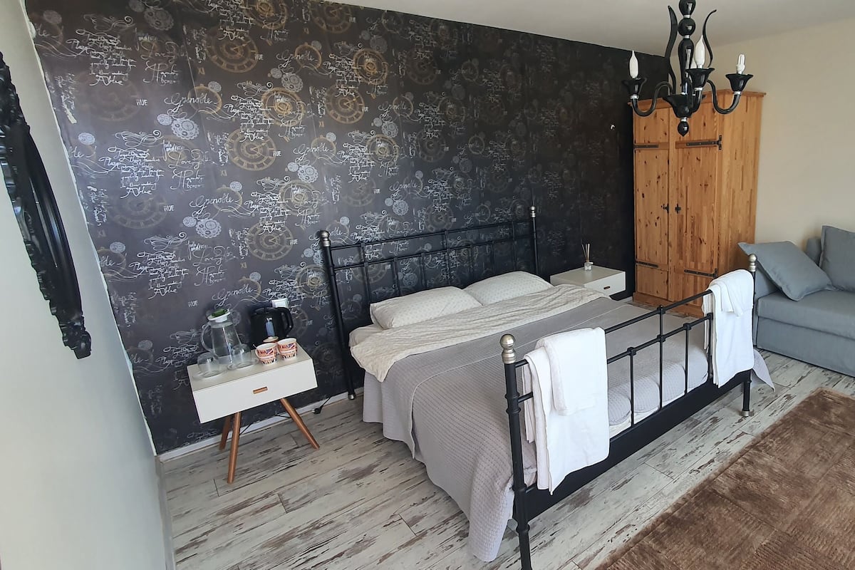 A bedroom with a bed, a grey sofa, a wooden wardrobe, a nightstand, and a wall with decorative patterns.