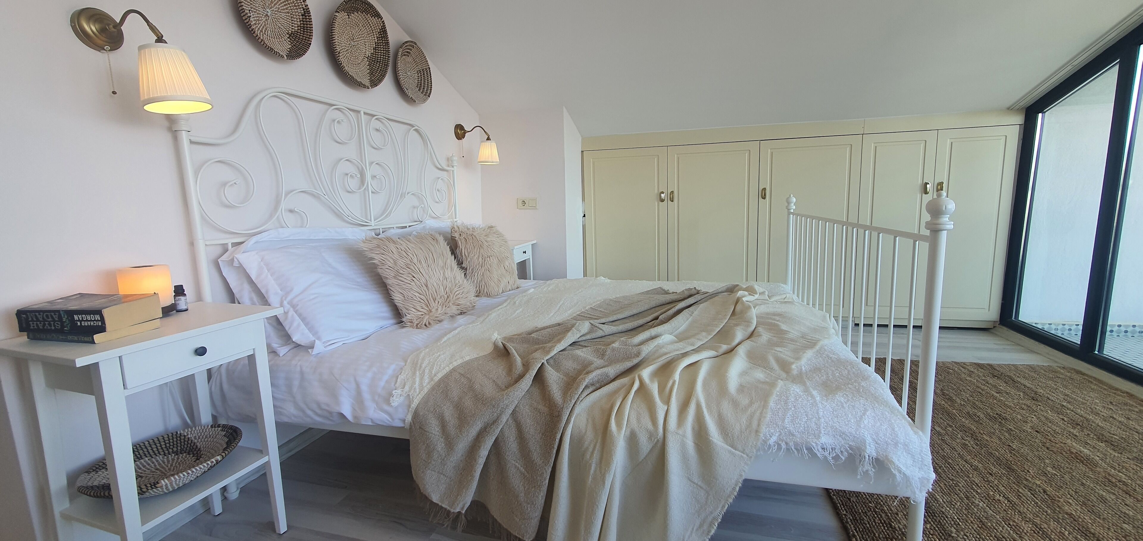 Un dormitorio con cama, mesitas de noche, lámpara, estantería y un amplio ventanal.