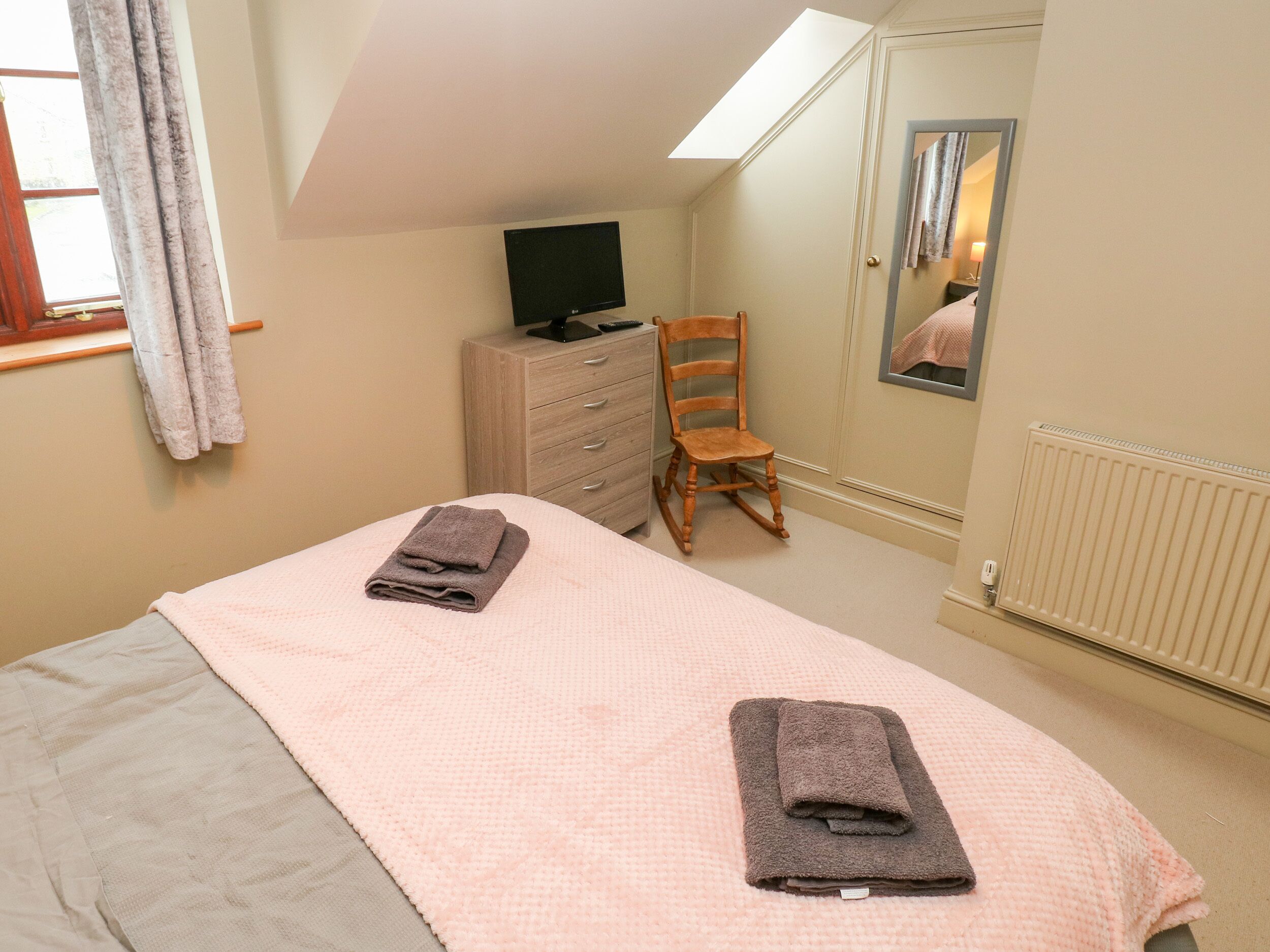 Un dormitorio con cama, cómoda, televisor, mecedora y ventana con cortinas.